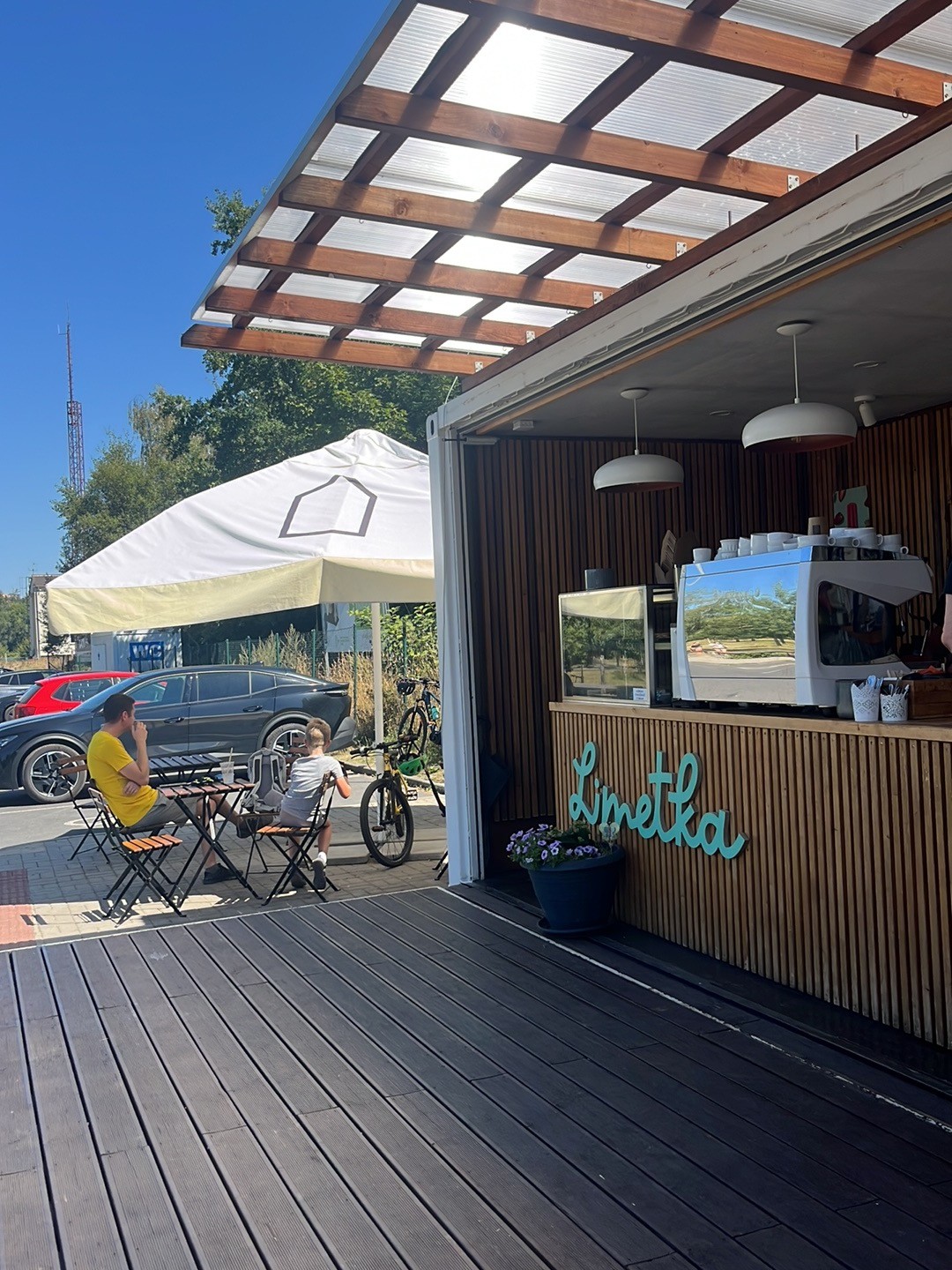 The Limetka v meandru shipping-container kiosk with its wood-slat counter and signage, seen from the outdoor terrace with parasol tables on a sunny day